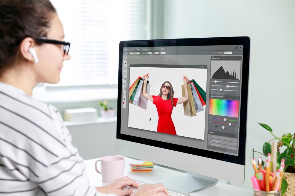 A woman wearing spectacles, listening to music on wireless earphones, seated at a desk in front of a computer, with a photo editor displayed on the screen.