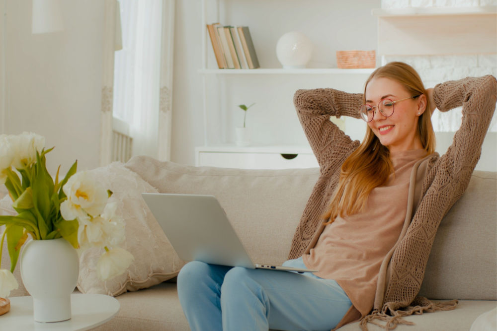 A woman sitting comfortably on her living room couch, with her hands resting behind her head, as she smiles at something on her laptop screen.