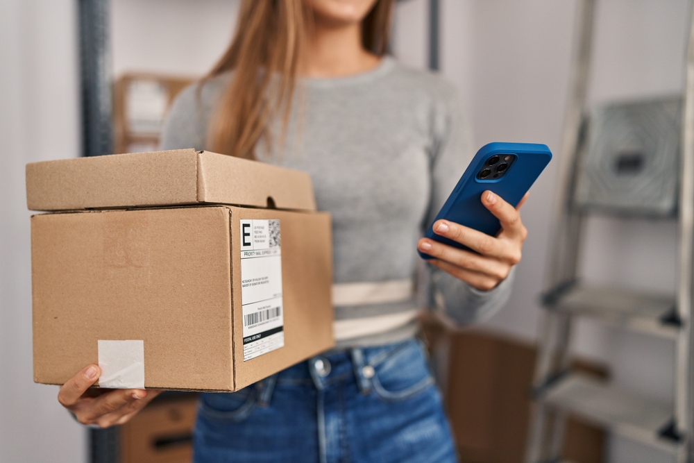 Young professional consulting her phone while getting boxes ready for shipping