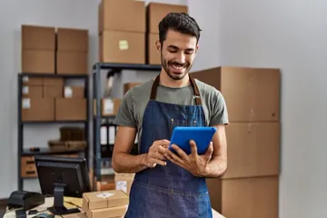 A man smiling, working on a table as he stands in his office, which is filled with shelves and boxes