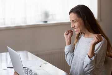 A young woman with long brown hair, wearing a light-grey jumper with the sleeves slightly rolled up, sitting at a desk in front of her laptop. She is seemingly ecstatic, with her arms lift, her hands in balls, and a huge smile on her face. On the table is a pile of papers. The interior of the room is white, with a big window faded out in the background.