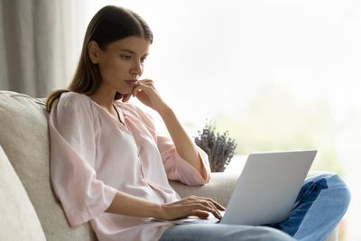 A young women with long brown hair, wearing a pink blouse and multi-shade denim jean, sitting cross-legged on a light grey couch. Her laptop is open on her lap, with her one arm propped op on the couch and her chin resting on this hand, and the other hand on the trackpad of the laptop. She has a neutral expression on her face. In the background, a large window, heavy grey curtains, and a bouquet of lavender are faded out.