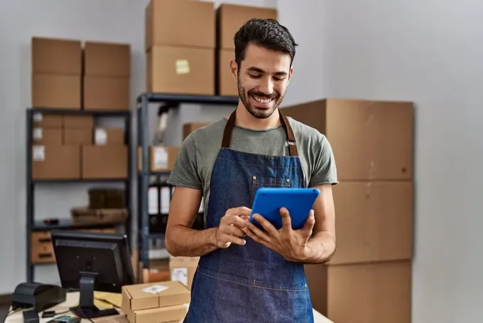 A man smiling, working on a table as he stands in his office, which is filled with shelves and boxes