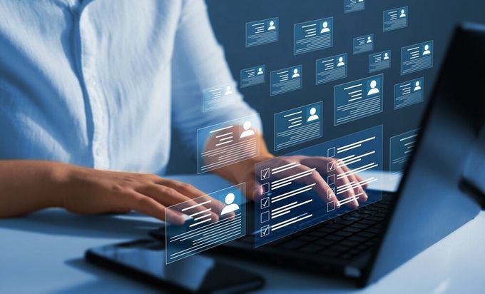 A person's torso, wearing a light blue long-sleeve button-up dress shirt, with the sleeves slightly rolled up. Both hands are typing on a black laptop, which is place atop a white coffee table. There is a black cellphone next to the laptop. There are multiple icon boxes featuring contact icons and task checklists hovering above the laptop's keyboard.