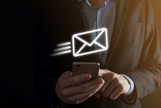 The torso of a man, wearing a blue button-up dress shirt and black blazer, standing with his phone in his one hand, typing with the other. On the right-hand corner of the image, there is an influx of warm light, whereas the left-hand side is black. In the middle of the image, we see an icon of an envelope, with three lines on the left of it.