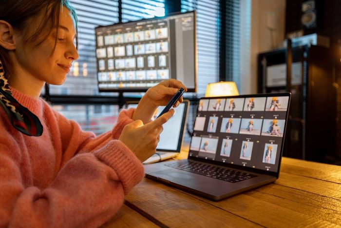 A woman seated at a desk, busy on her phone, with a laptop and a secondary screen on the table, with various images on display.