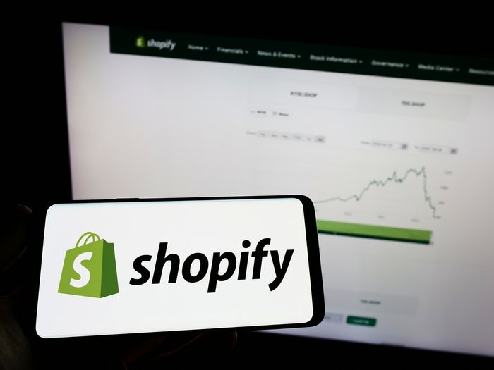 Mobile phone displaying the Shopify logo with a computer screen in the background showing a graph